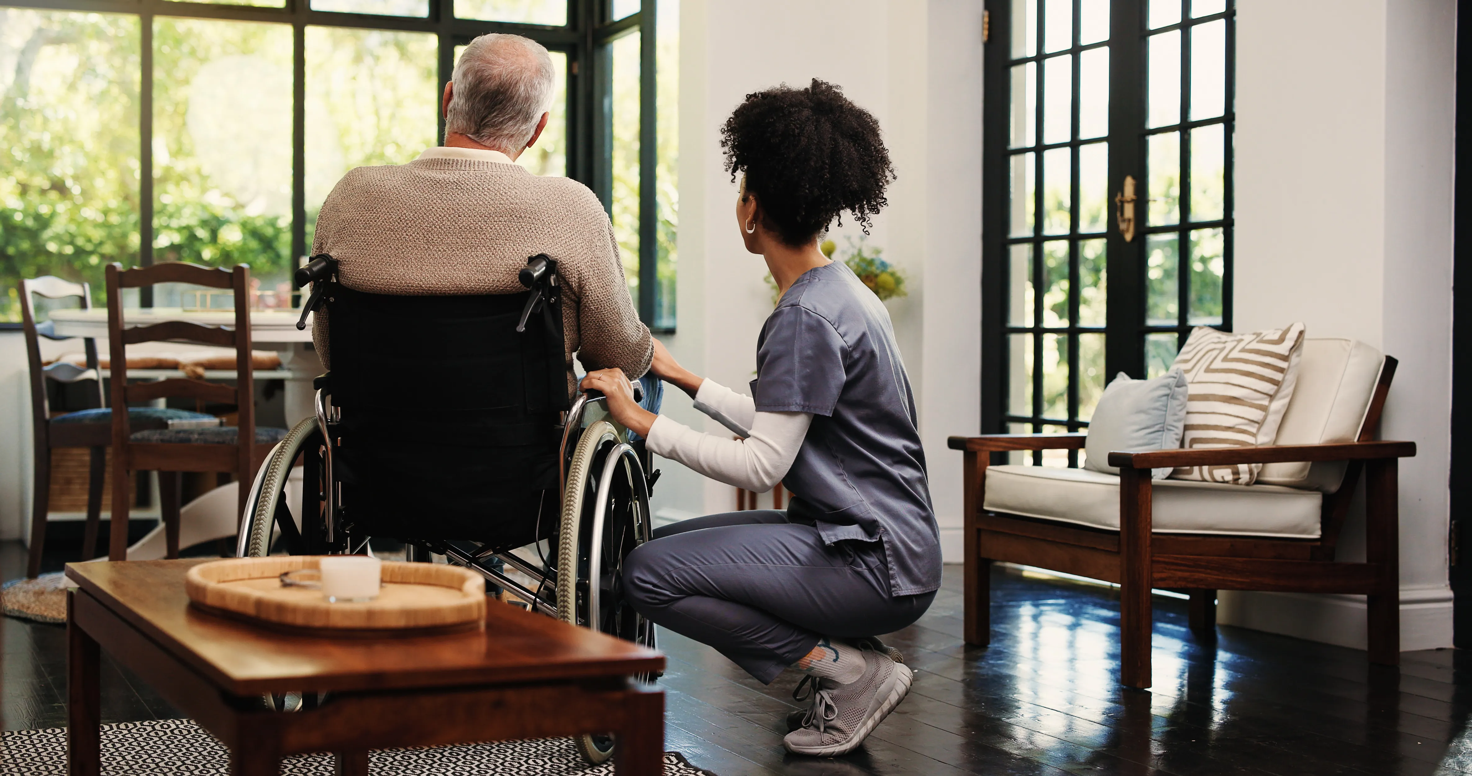 Morning Star caregiver supports a senior man in a wheelchair beside bright windows in his cozy home.